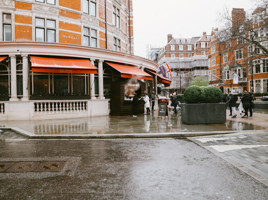 A wet pavement outside a building with an orange awning, featuring a sidewalk café with a smoking chimney on the corner. Several people are walking or standing nearby in coats, indicating cool weathe